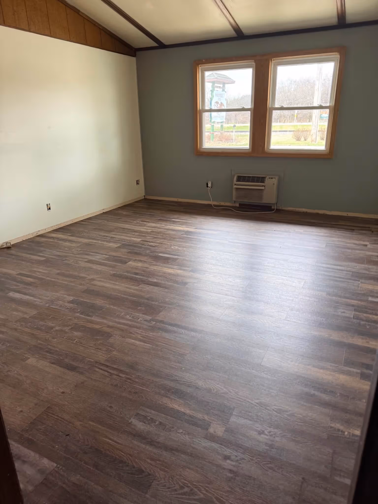 Empty room with wood-patterned floor, pale blue and white walls, two windows, and a wall air conditioner.