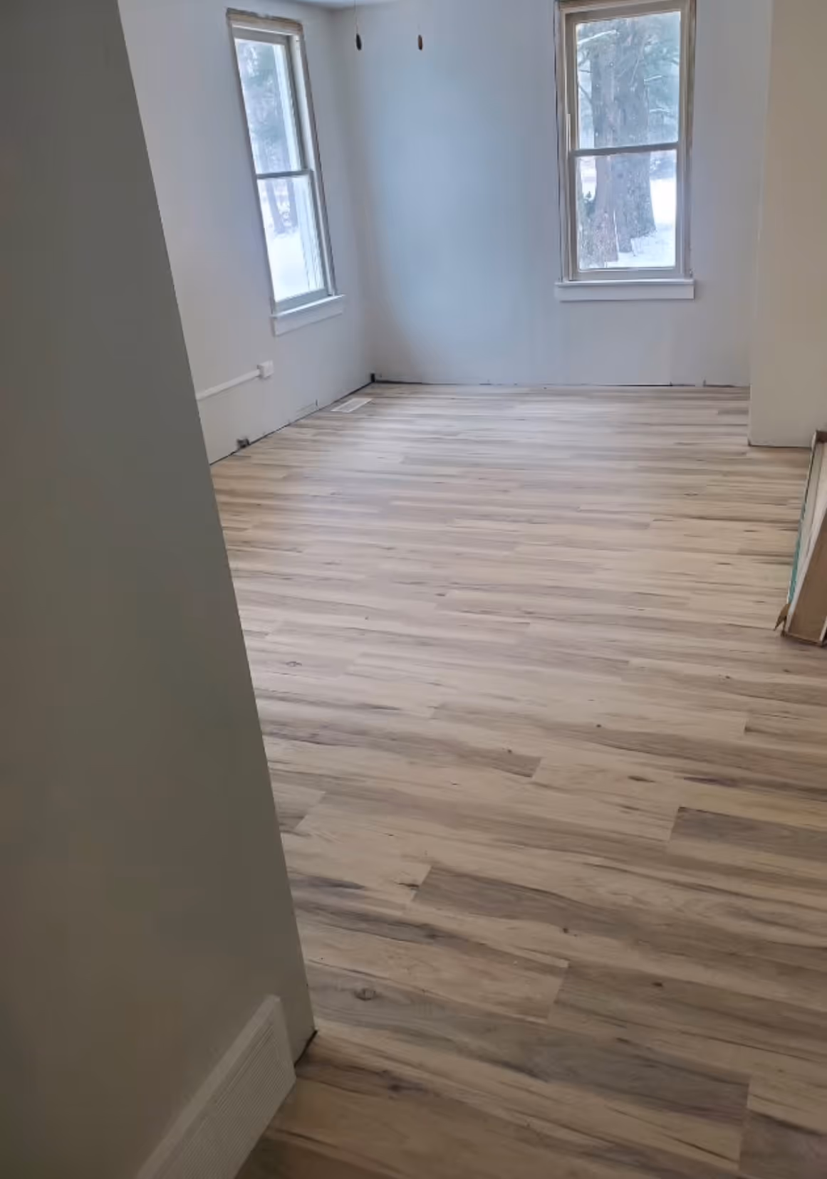Empty room with light wood laminate flooring and two large windows showing snowy trees outside.