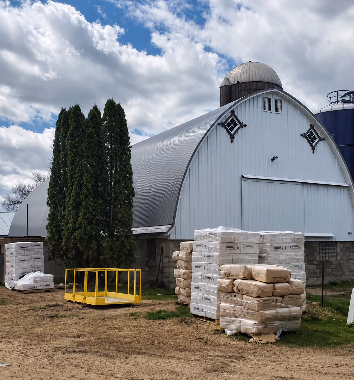 White barn with silver curved roof, silo, trees, and pallets of wrapped building materials outside.
