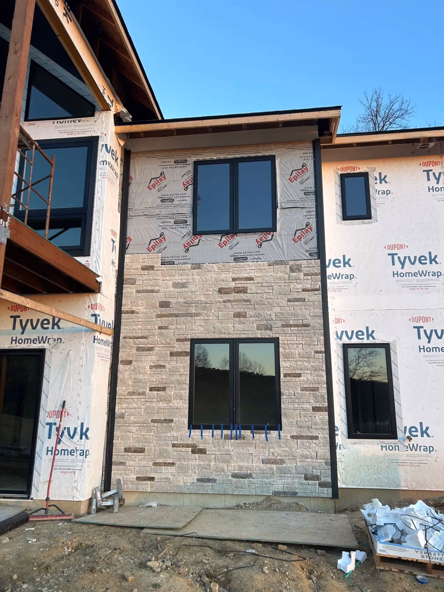 Partially constructed house with stone siding on the ground floor and windows reflecting trees.
