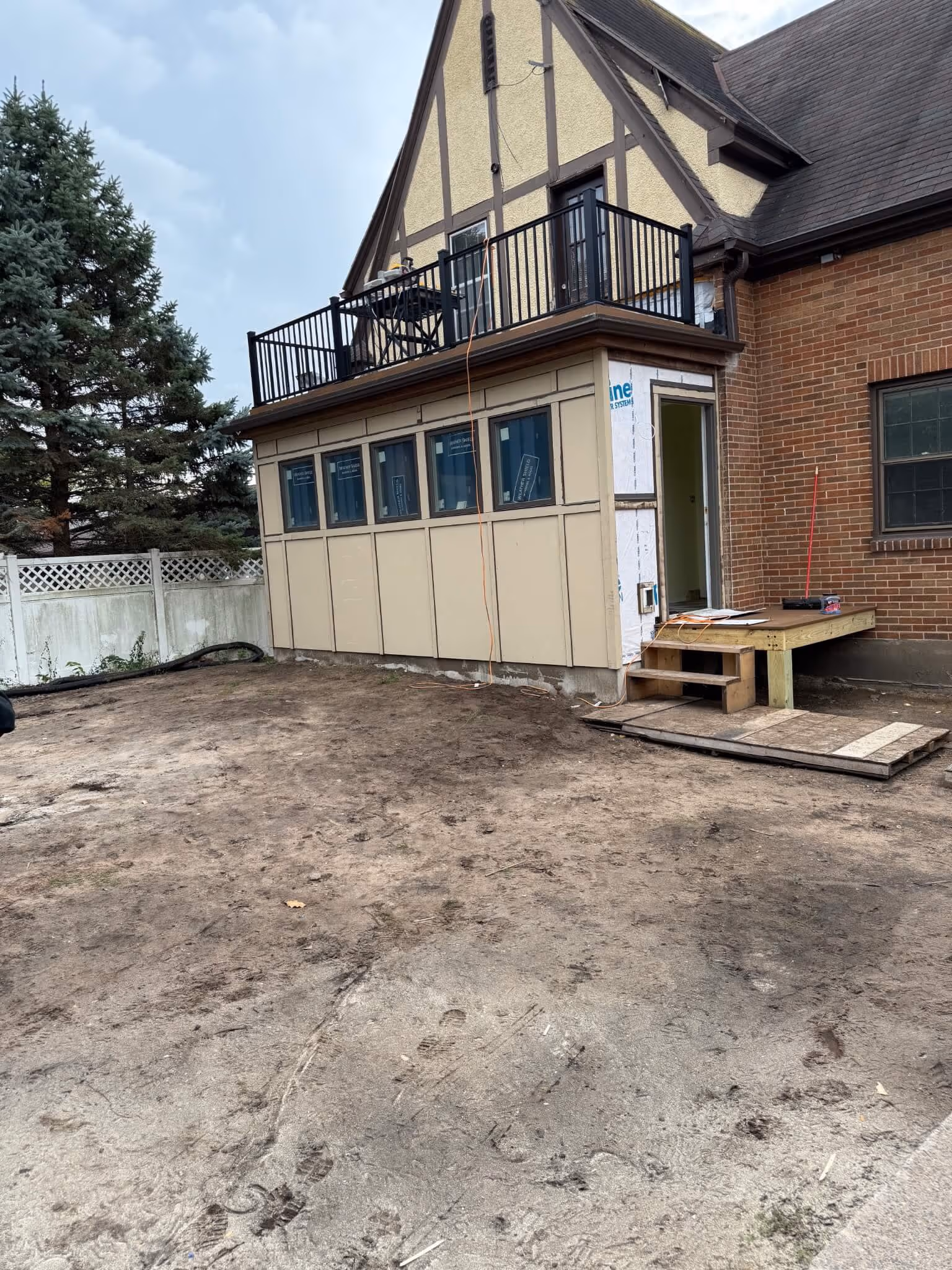 Backyard with dirt ground and a house extension featuring multiple windows and a balcony above.