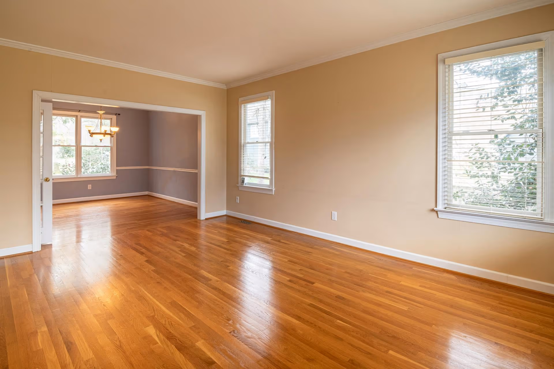Empty room with polished hardwood floor, beige walls, and two windows with blinds.